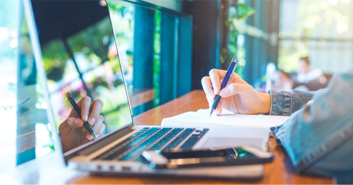 Person writing on a notebook in front of an open laptop