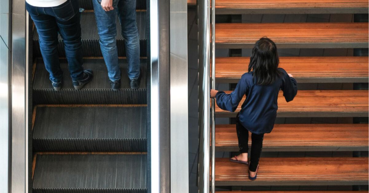 Two men taking the escalator to go up while a woman climbs stairs, next to the escalator, to go up