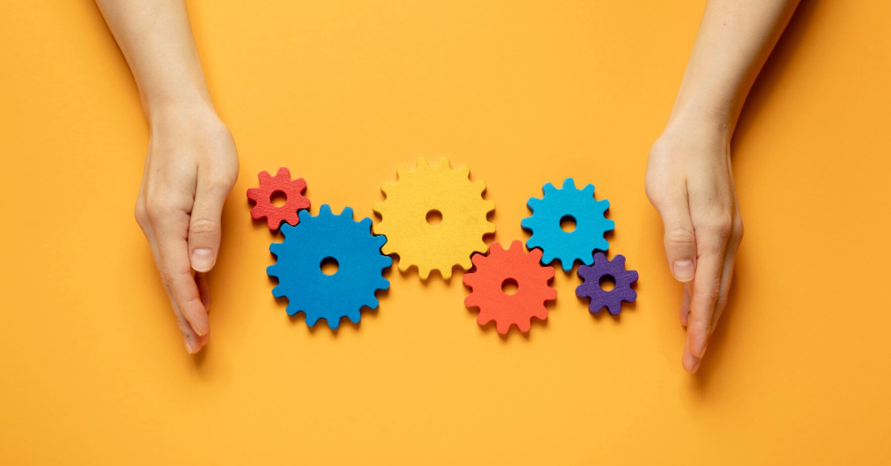 Hands protecting multiple coloured wooden gears against a yellow background