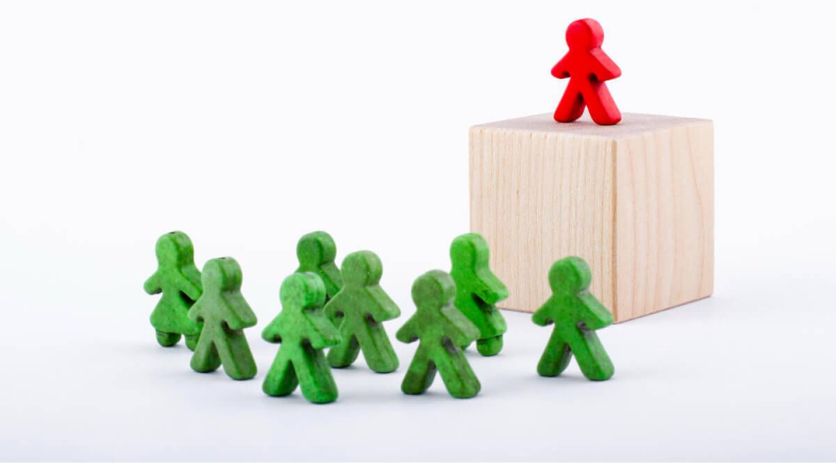 Red wooden human on top of a wooden block looking down on green wooden humans